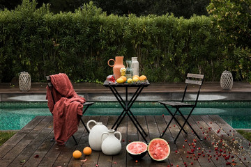 Diseño de exteriores Diseño de exterior, en frente de una piscina, con vegetación y una mesa y sillas de exterior. Sobre la mesa y en el suelo, distintos elementos como jaras y fruta.