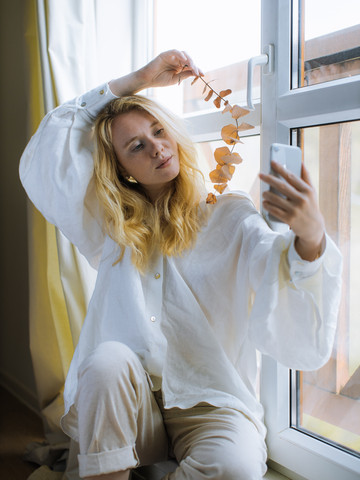 Chica frente a una ventana fotografiando con el móvil unas hojas